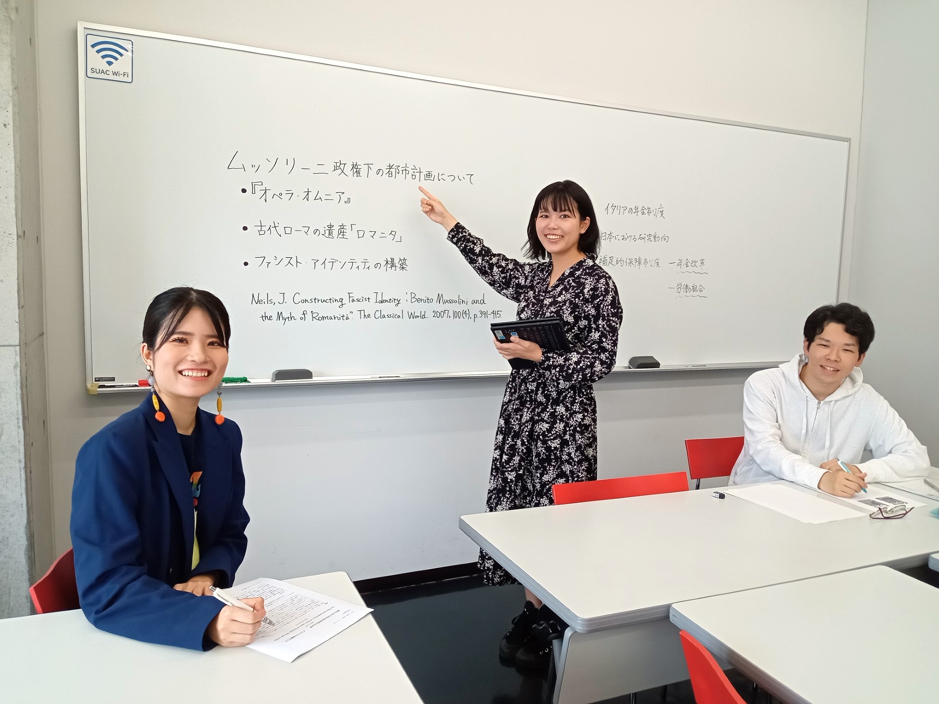ゼミの様子（ホワイトボードの前で指さす学生とそれを聞いている2名の学生）